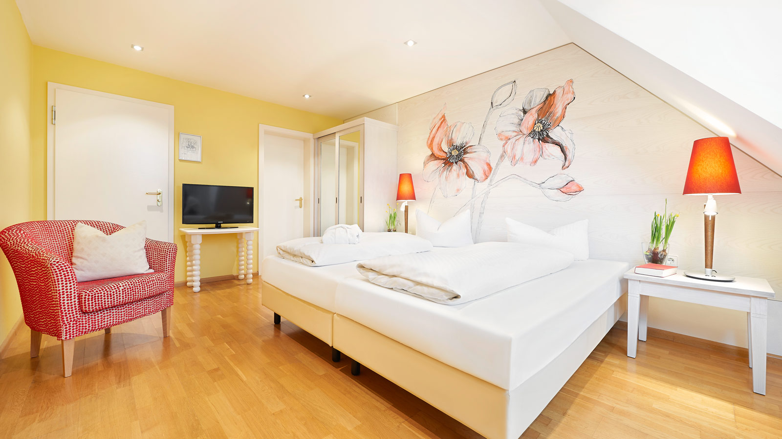 Bright and welcoming hotel room with a large double bed, floral wall artwork, wooden flooring and a comfortable armchair.