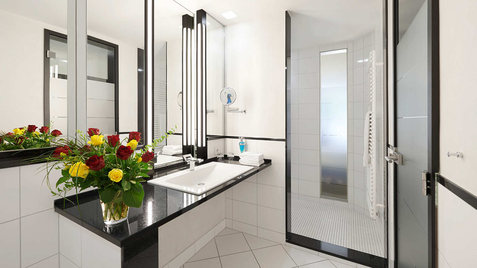 Elegantes Badezimmer mit großzügigem Waschtisch, beleuchtetem Spiegel, frischem Blumenstrauß und bodengleicher Dusche.