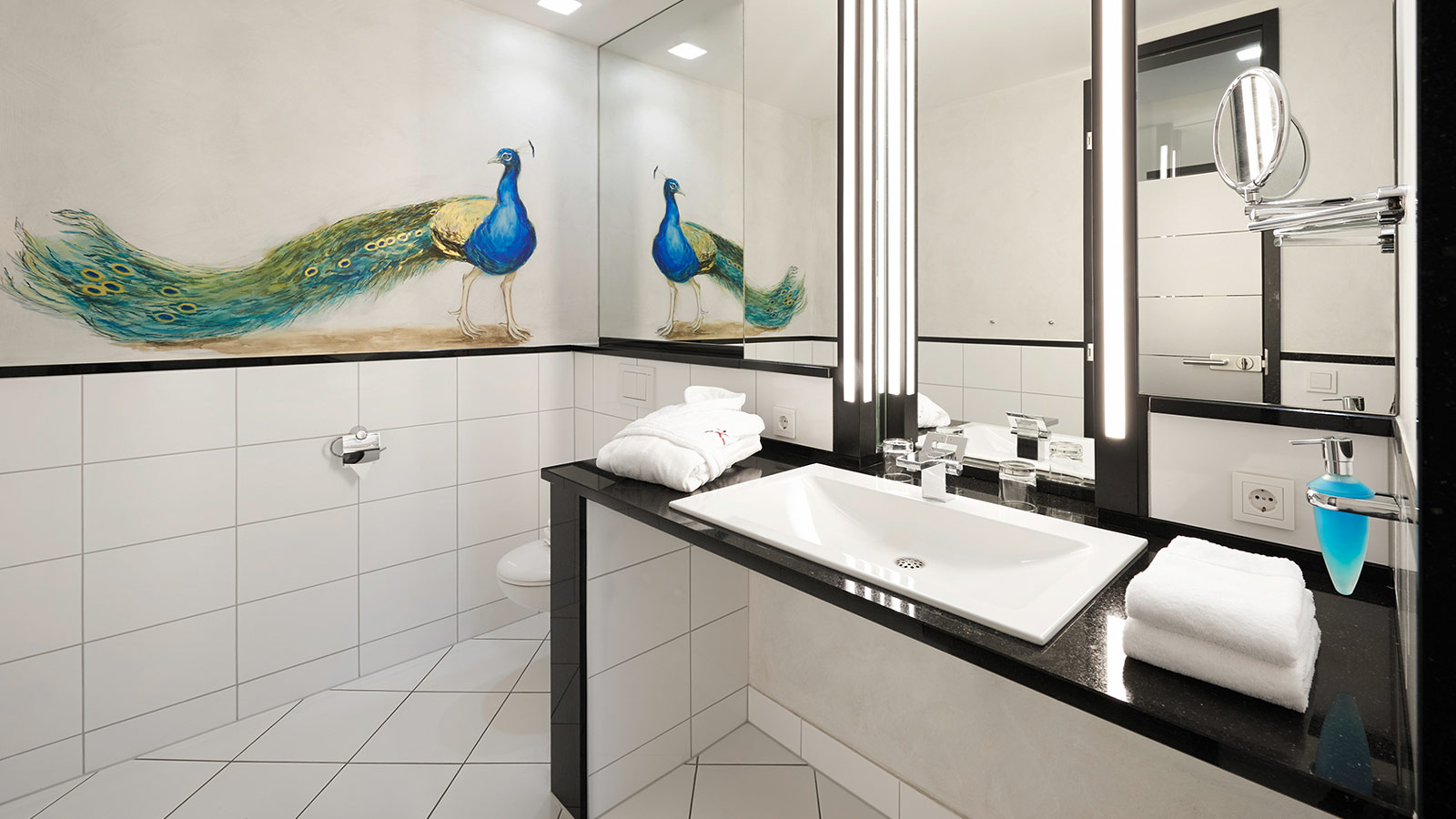Modernes Badezimmer mit beleuchtetem Spiegel, großem Waschtisch, Pfauenmotiv an der Wand und hochwertiger Ausstattung.